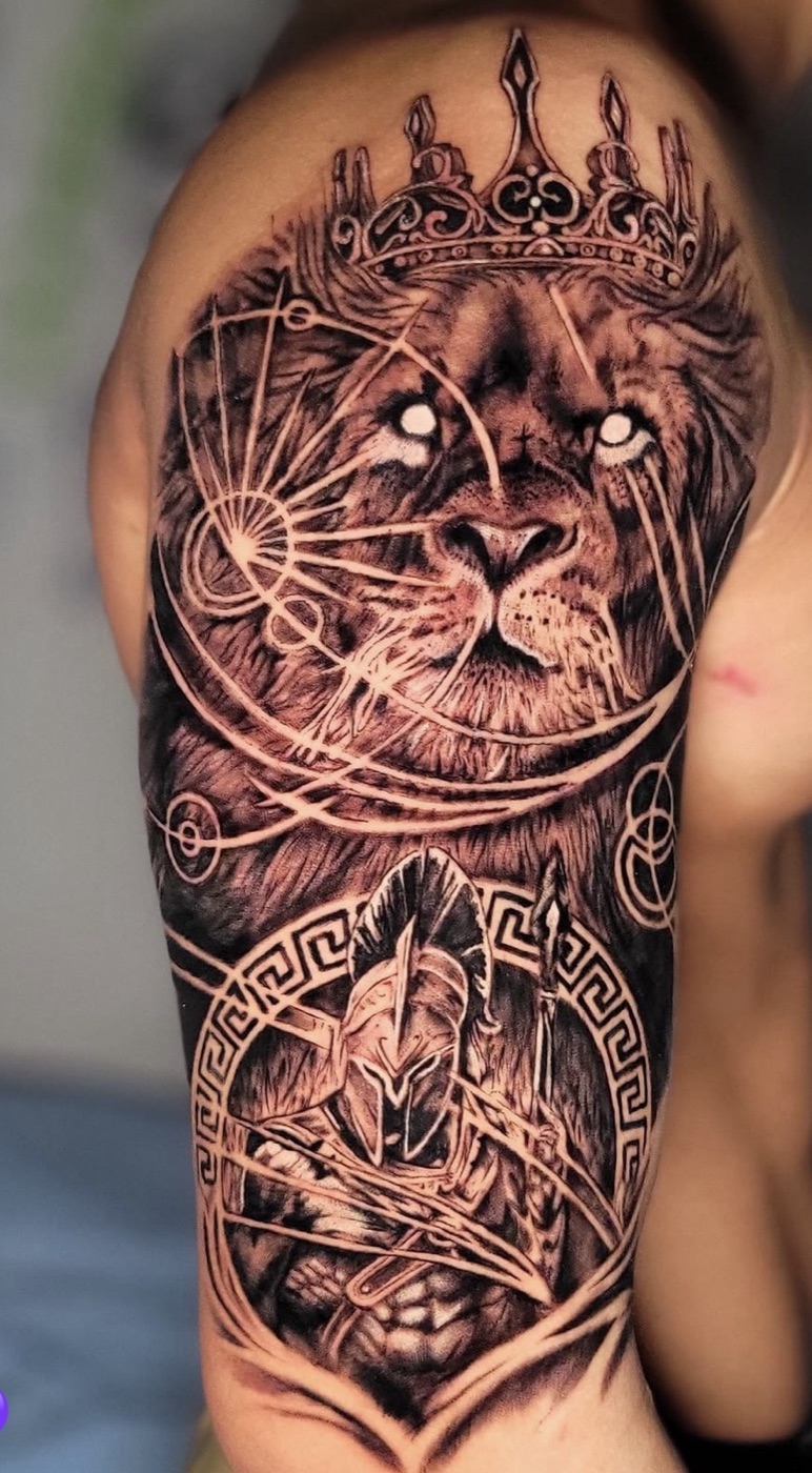 Black and grey realism tattoo of a crowned lion over a Spartan warrior, full upper arm